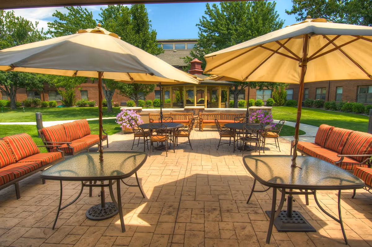 Outdoor patio area with glass tables, striped cushioned benches, and large beige umbrellas providing shade. In the background, there is a gazebo surrounded by green grass, trees, and a brick building.