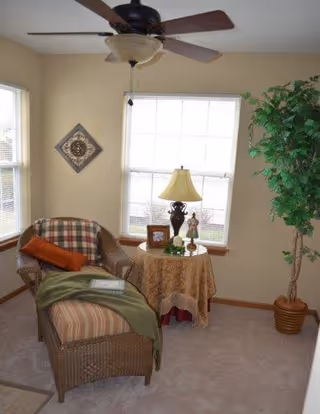 A cozy corner of a living room with a wicker chaise lounge covered with a striped cushion, a green throw blanket, and an orange pillow. Next to it is a round table draped with a patterned cloth, holding a decorative lamp, a small framed photo, and a figurine. A large potted plant stands to the right, and two windows with blinds let in natural light. A ceiling fan with a light fixture hangs above.