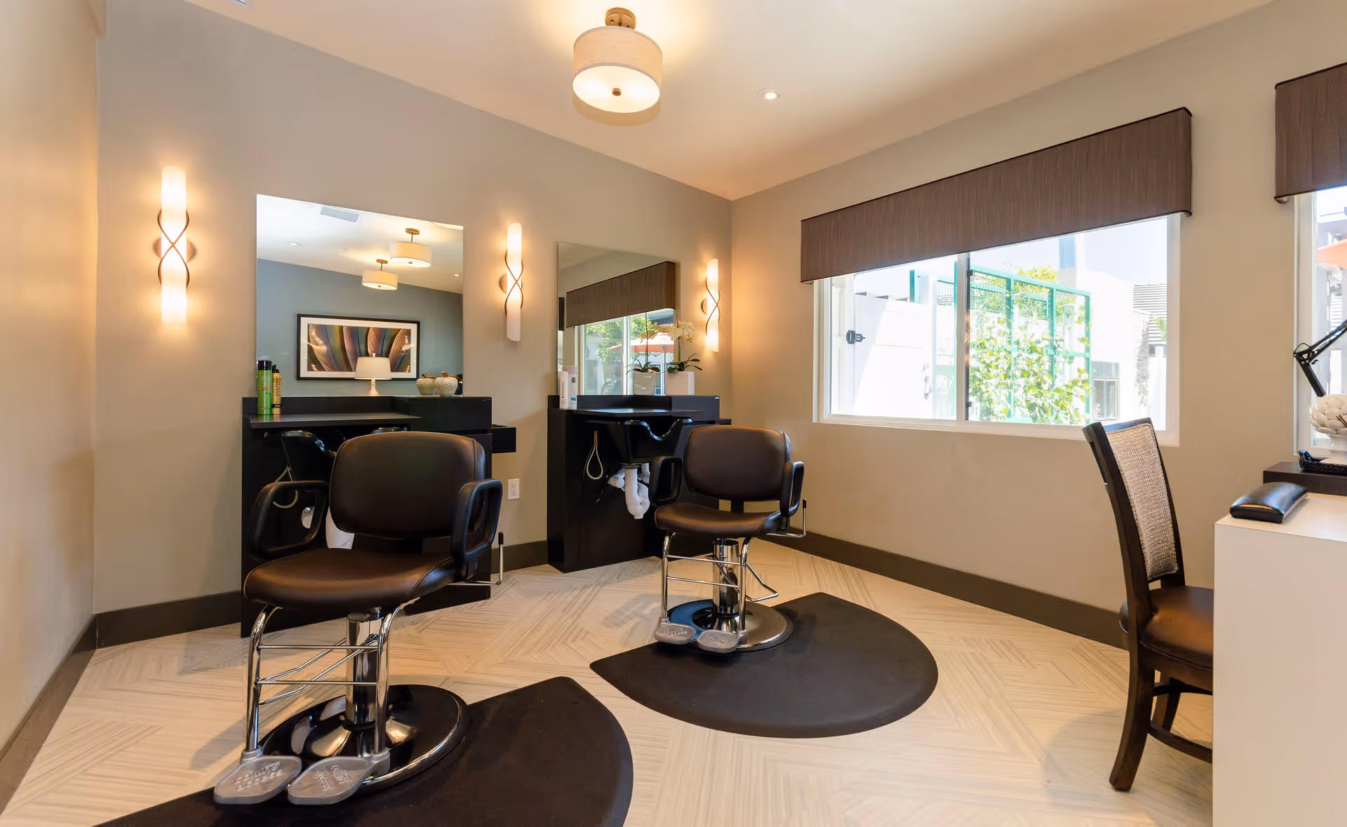 Interior of a modern salon area with two black salon chairs in front of mirrors and sinks, soft wall lighting, a large window with a brown valance, and a chair next to a desk on the right side.