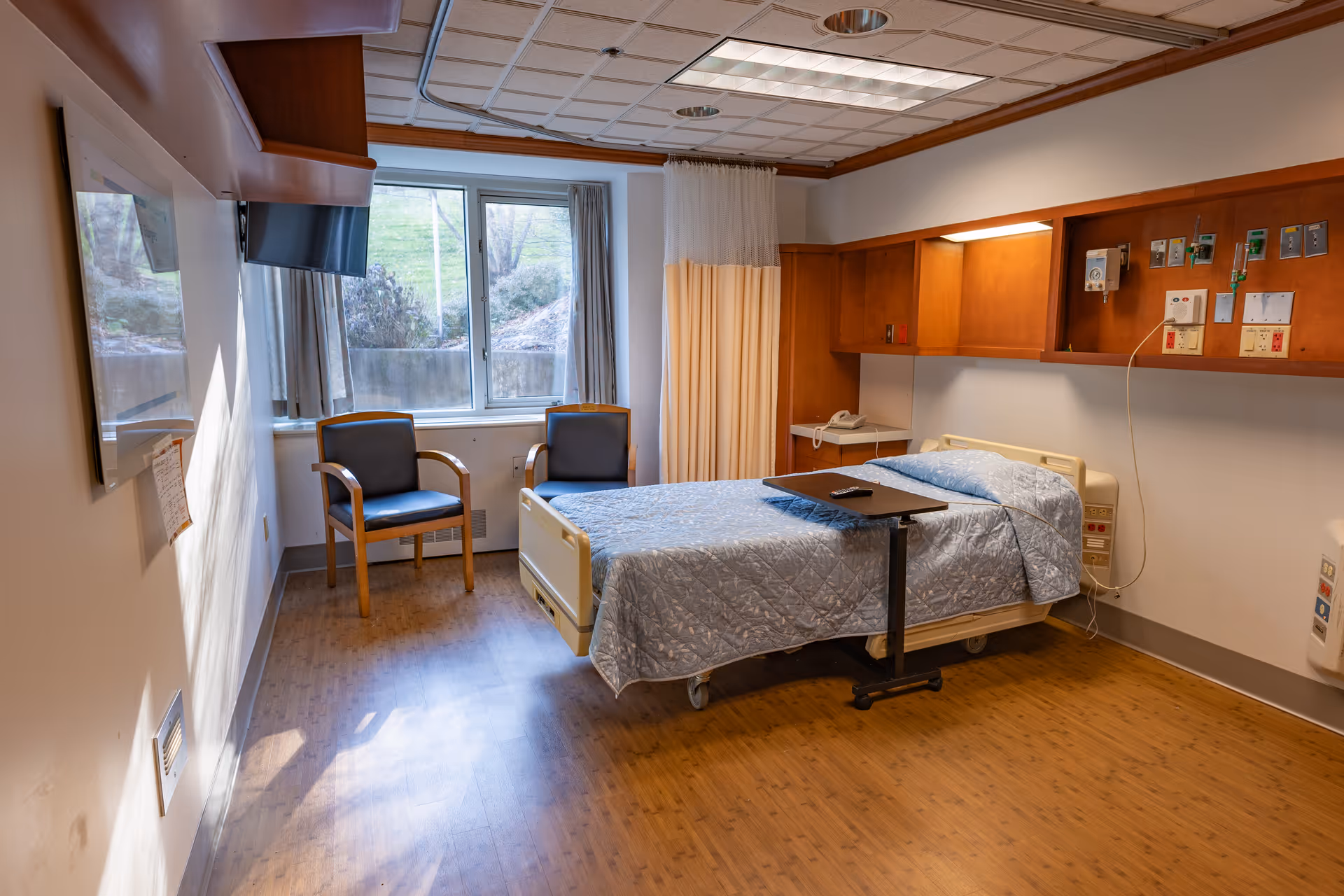A hospital-style patient room with a single bed covered in a blue quilted blanket, a small overbed table with a remote control on it, two chairs near a window with curtains, a wall-mounted TV, and medical equipment mounted on the wall above the bed. The room has wood flooring and a ceiling with recessed lighting.