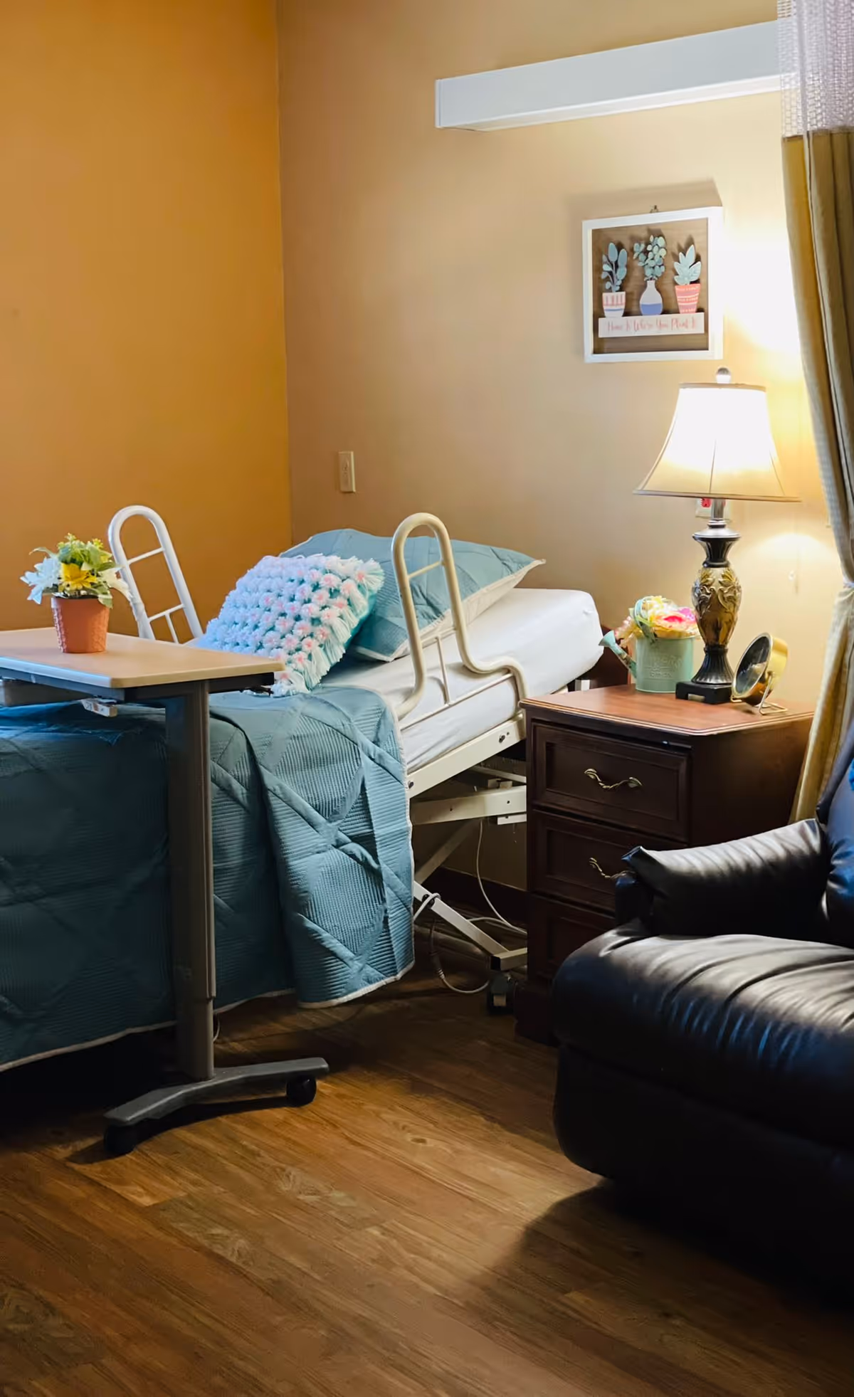 A tidy patient bedroom with an adjustable hospital bed covered in teal bedding, a bedside table with a lamp and flowers, an overbed table, and a leather recliner.