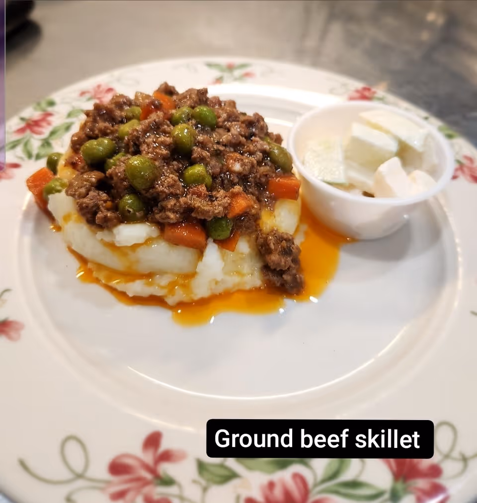A plate with a serving of ground beef skillet containing ground beef, peas, and diced carrots on a bed of mashed potatoes, accompanied by a small cup of sliced white cheese or vegetable on the side. The plate has a floral pattern around the rim.