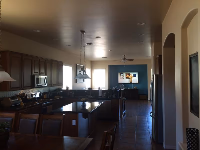 Interior view of a modern kitchen and dining area with dark wooden cabinets, a kitchen island with a glossy countertop, hanging pendant lights, and a dining table with chairs in the foreground. In the background, there is a living area with a blue accent wall, a ceiling fan, and a TV mounted on the wall.