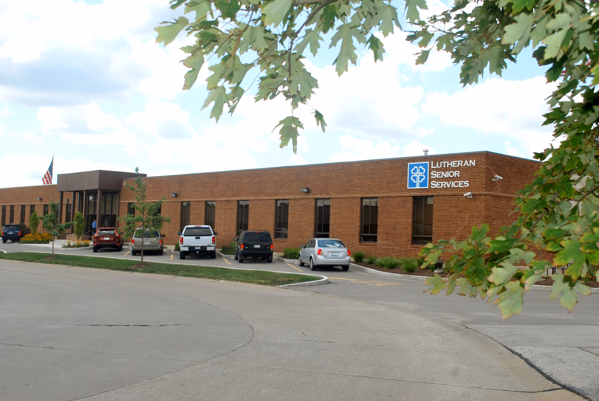 Exterior view of a single-story brick building with a sign that reads 'Lutheran Senior Services'. Several cars are parked in front of the building, and there are trees with green leaves partially framing the image. The sky is partly cloudy.