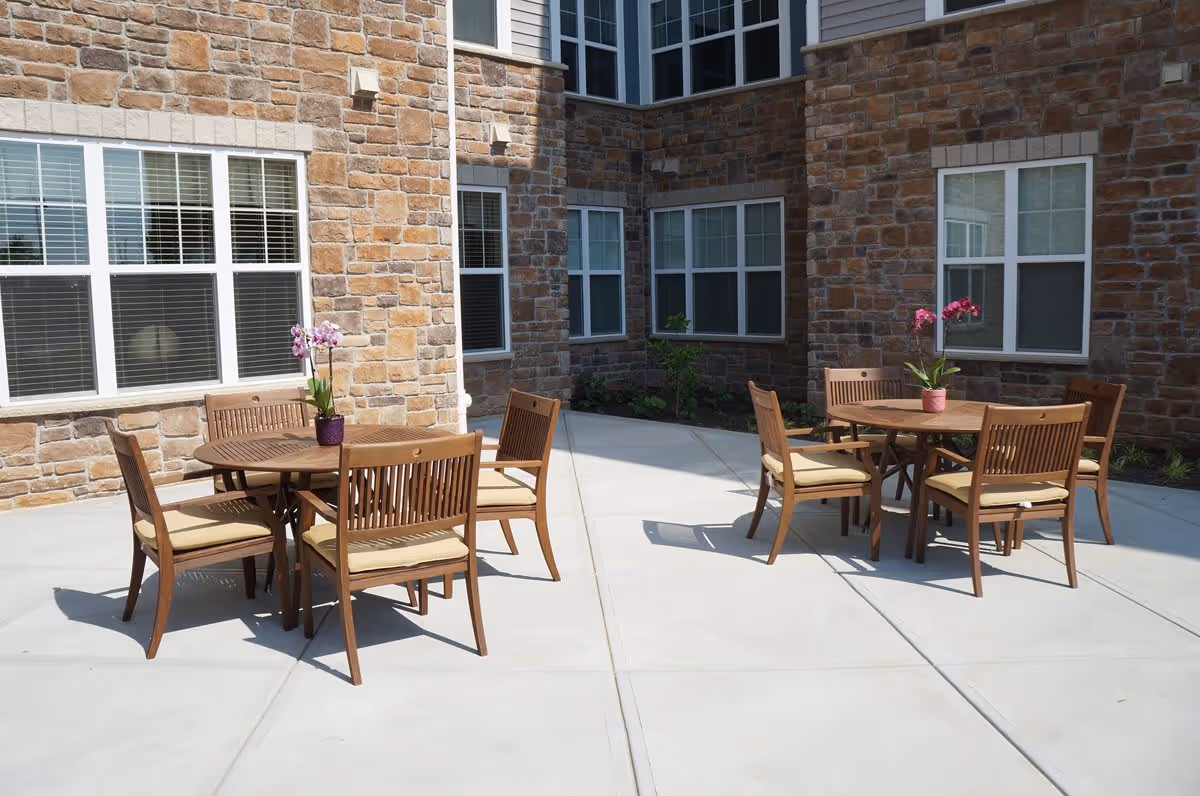 Outdoor patio area at Greens at English Consul featuring two round wooden tables each surrounded by four wooden chairs with beige cushions. Each table has a small potted orchid plant. The patio is paved with light-colored concrete and is adjacent to a building with stone and siding exterior walls and multiple windows.
