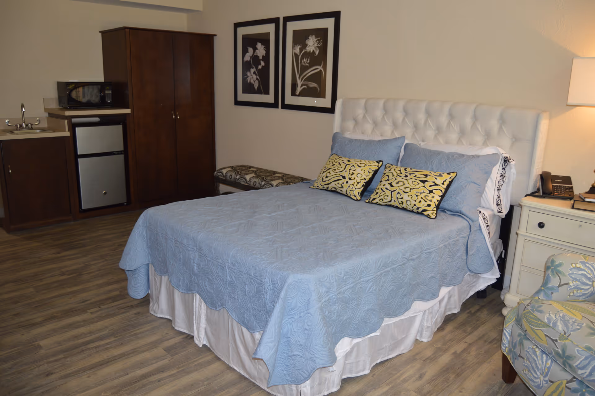 A senior living facility bedroom with a neatly made bed covered in a light blue quilt and decorative pillows. The room features a small kitchenette area with a sink, microwave, and mini fridge, a wooden wardrobe, a patterned bench, two framed floral artworks on the wall, a nightstand with a lamp and phone, and a floral upholstered chair. The floor is wood-style laminate.