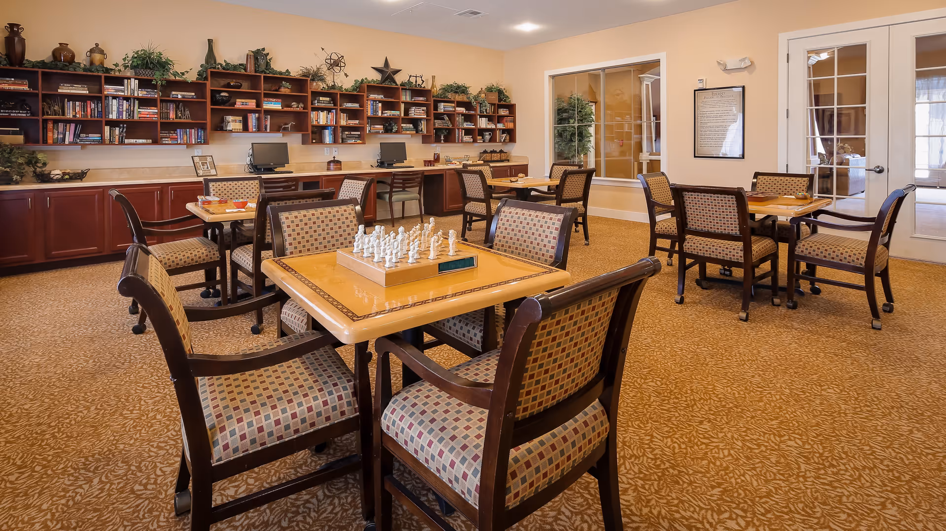 A well-lit activity room with multiple wooden tables and cushioned chairs arranged neatly. One table in the foreground has a chess set on it. The back wall features built-in wooden shelves filled with books, decorative items, and two desktop computers. The room has beige walls and carpeted flooring with a patterned design. There are large windows and a glass door on the right side, allowing natural light to enter.