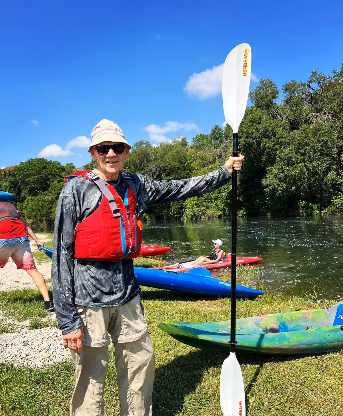 An elderly man wearing a red life jacket, beige pants, a light-colored hat, and sunglasses is standing on grass near a river, holding a white kayak paddle vertically. Behind him, there are colorful kayaks on the grass and a person sitting in a red kayak on the water. Trees and a clear blue sky with a few clouds are visible in the background.