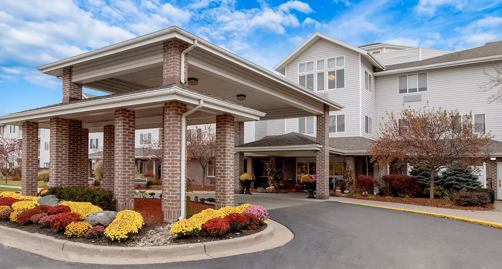 Entrance of a senior living facility with a covered drop-off area supported by brick columns, surrounded by landscaped flower beds with yellow and red flowers, and a multi-story building in the background under a partly cloudy sky.