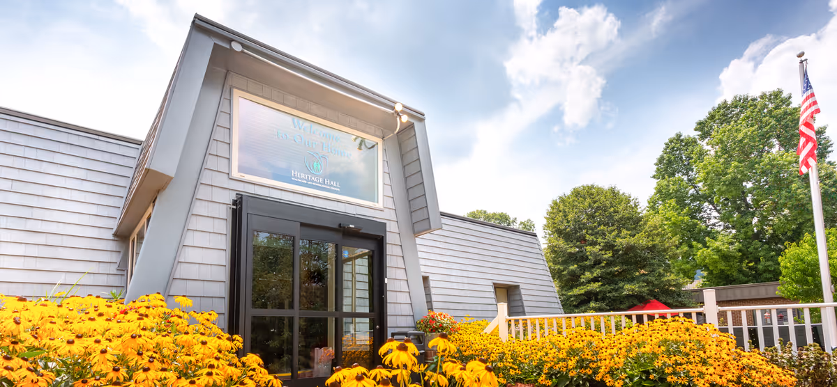 Exterior front entrance of Heritage Hall Rich Creek facility with a sign above the door that reads 'Welcome to Our Home Heritage Hall'. The building has gray siding and is surrounded by vibrant yellow flowers and green trees under a partly cloudy sky. An American flag is visible on the right side.