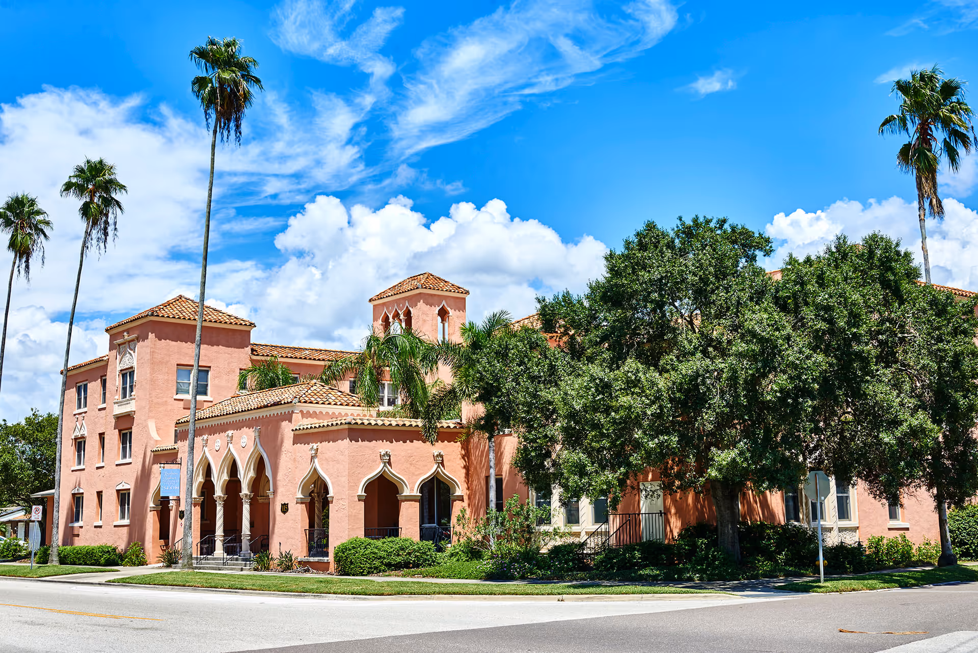 Exterior view of Hudson Manor Assisted Living, a pink stucco building with Spanish-style architecture, including arched windows and tiled roofs, surrounded by palm trees and greenery under a bright blue sky with scattered clouds.