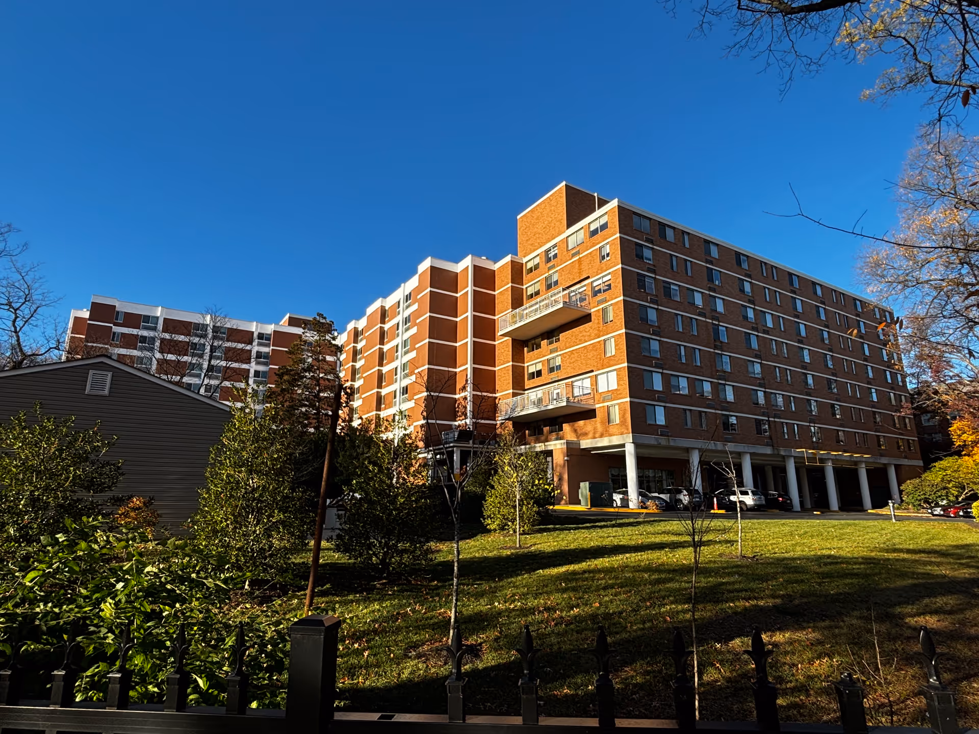 Exterior view of a multi-story brick building with balconies and numerous windows, surrounded by trees and greenery under a clear blue sky.