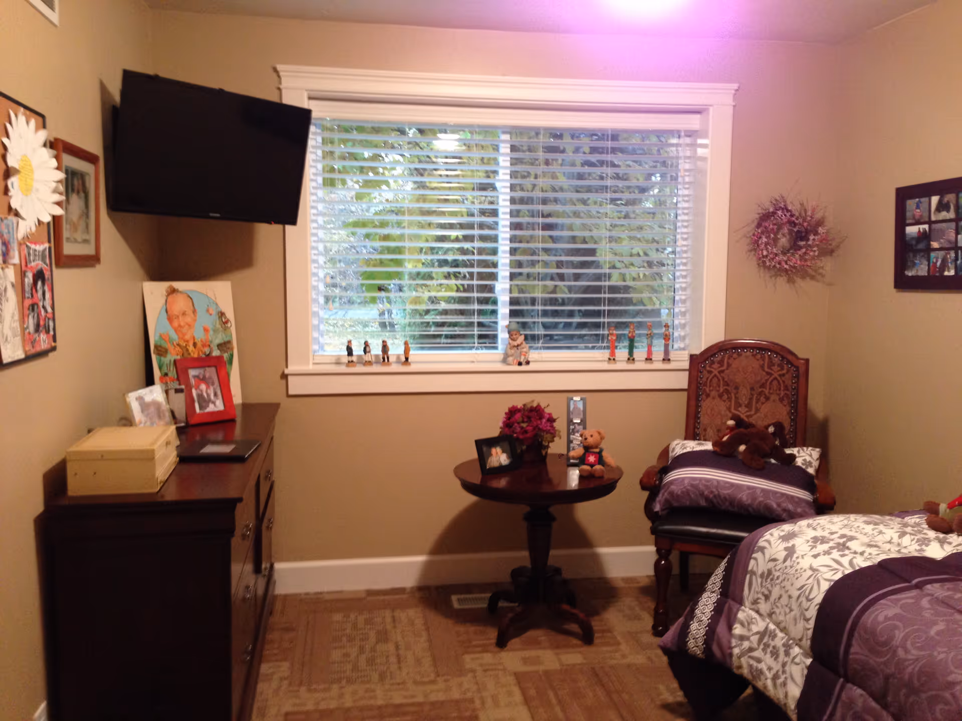 A cozy bedroom with a bed covered in purple and white patterned bedding, a wooden chair with pillows and stuffed animals, a small round table with a framed photo, flowers, and a teddy bear, a dresser with framed pictures and decorations, a wall-mounted TV, and a window with white blinds showing greenery outside.