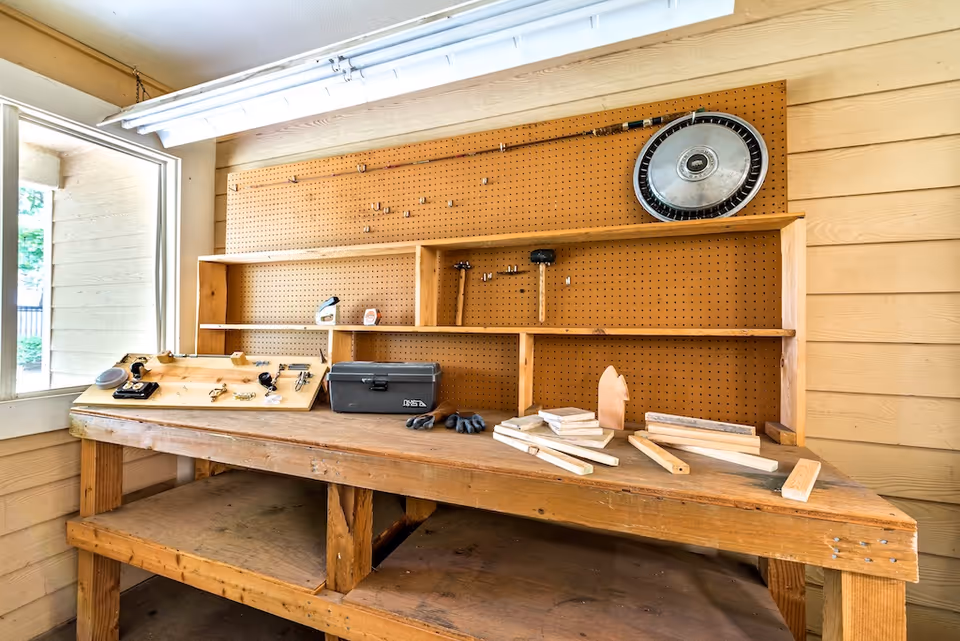 A wooden workbench with various tools and wooden pieces on it, set against a pegboard wall with a fishing rod and a hubcap hanging. The workbench is located near a window with natural light coming in.