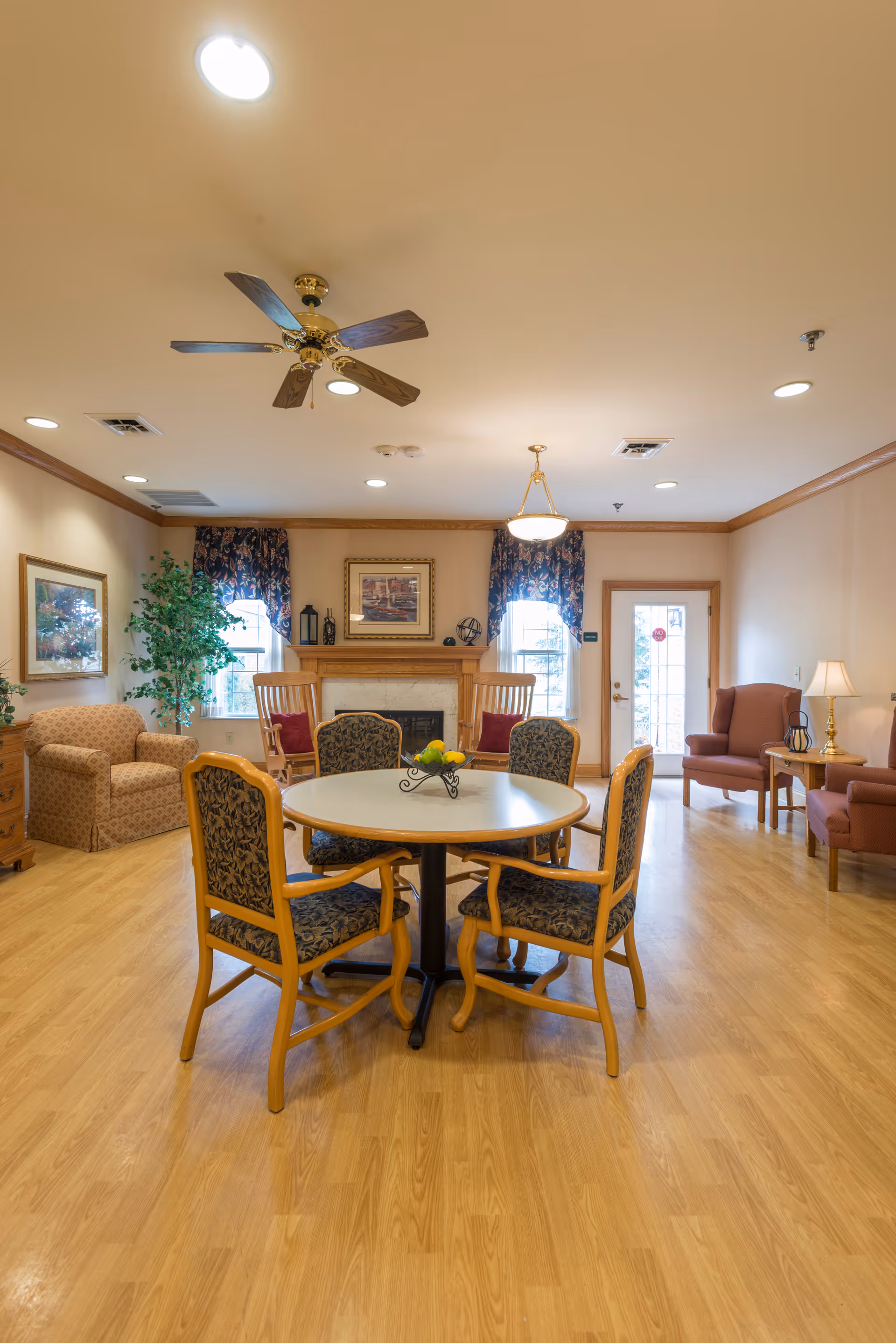 A bright communal lounge with a round table and four chairs in the center, armchairs, a fireplace, and windows in the background.