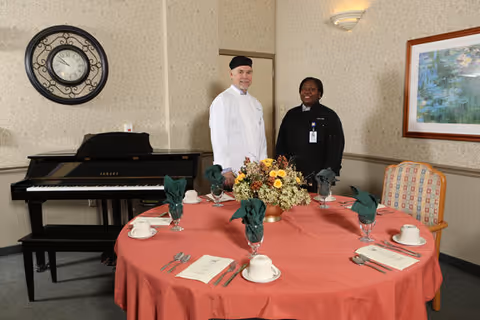 A round dining table set with place settings and a floral centerpiece, with two staff members standing behind it in a dining area featuring a piano and wall clock.