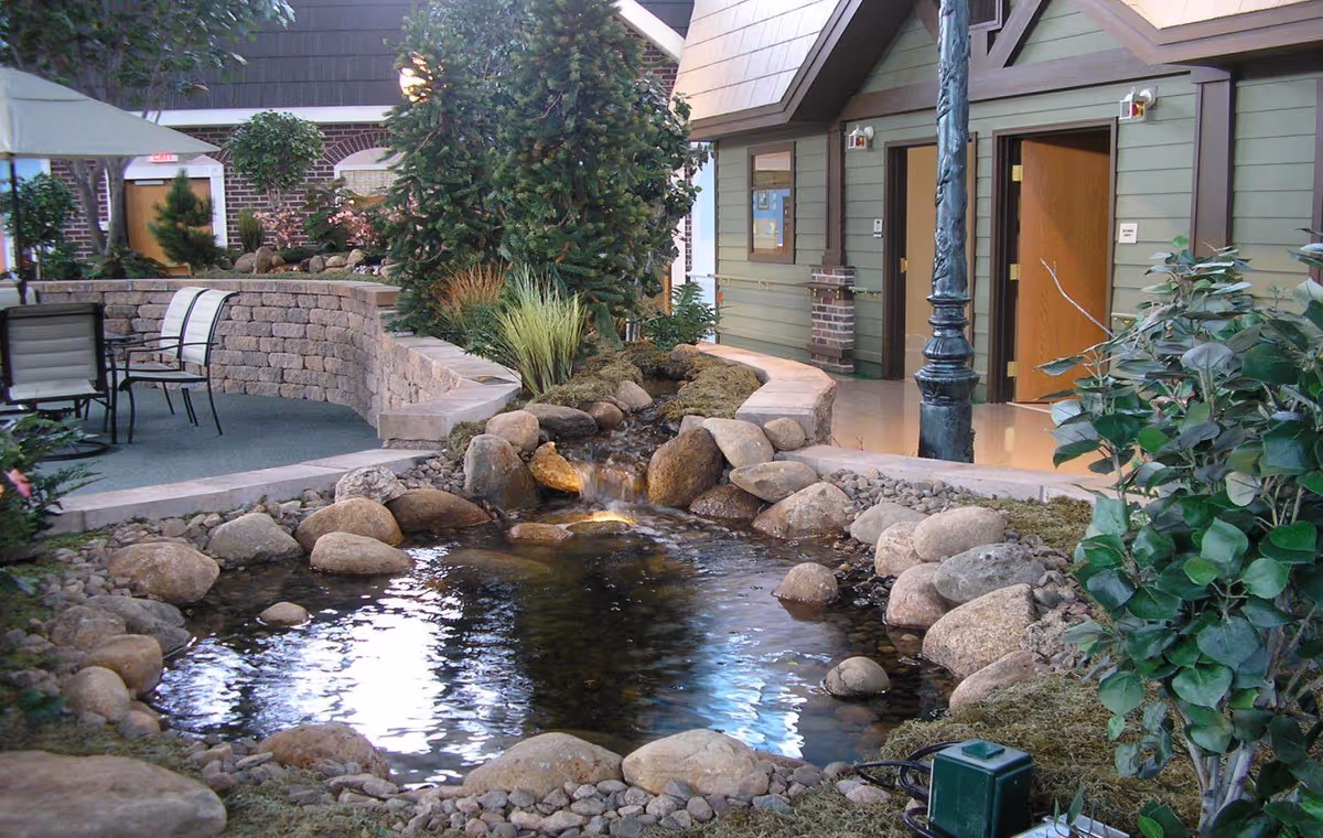 Indoor garden area with a small pond surrounded by rocks and plants. There is a curved stone bench and patio chairs on the left side, and a green building wall with open doors on the right. A black lamppost stands near the pond.