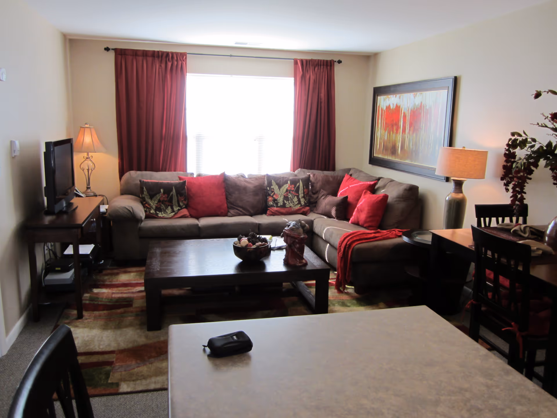 A cozy living room with a large L-shaped brown sofa adorned with red and patterned cushions. There is a dark wooden coffee table in front of the sofa with decorative items on it. The room has a window with red curtains, a floor lamp on the left side, and a table lamp on the right side next to a dining table with chairs. A colorful abstract painting hangs on the wall above the sofa.