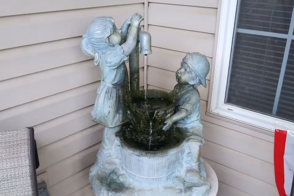 A decorative water fountain featuring statues of a girl drinking from a faucet and a boy sitting beside a bucket, placed on a round table against the exterior siding of a building near a window.