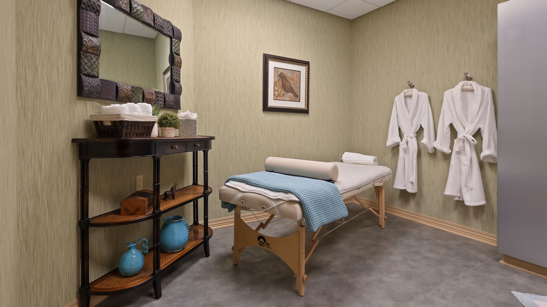 A serene massage room with a massage table covered with a white sheet and a blue blanket. Two white bathrobes hang on the wall. A black console table with shelves holds folded towels, a small plant, and decorative blue vases. A framed picture of a bird hangs on the wall above the massage table.