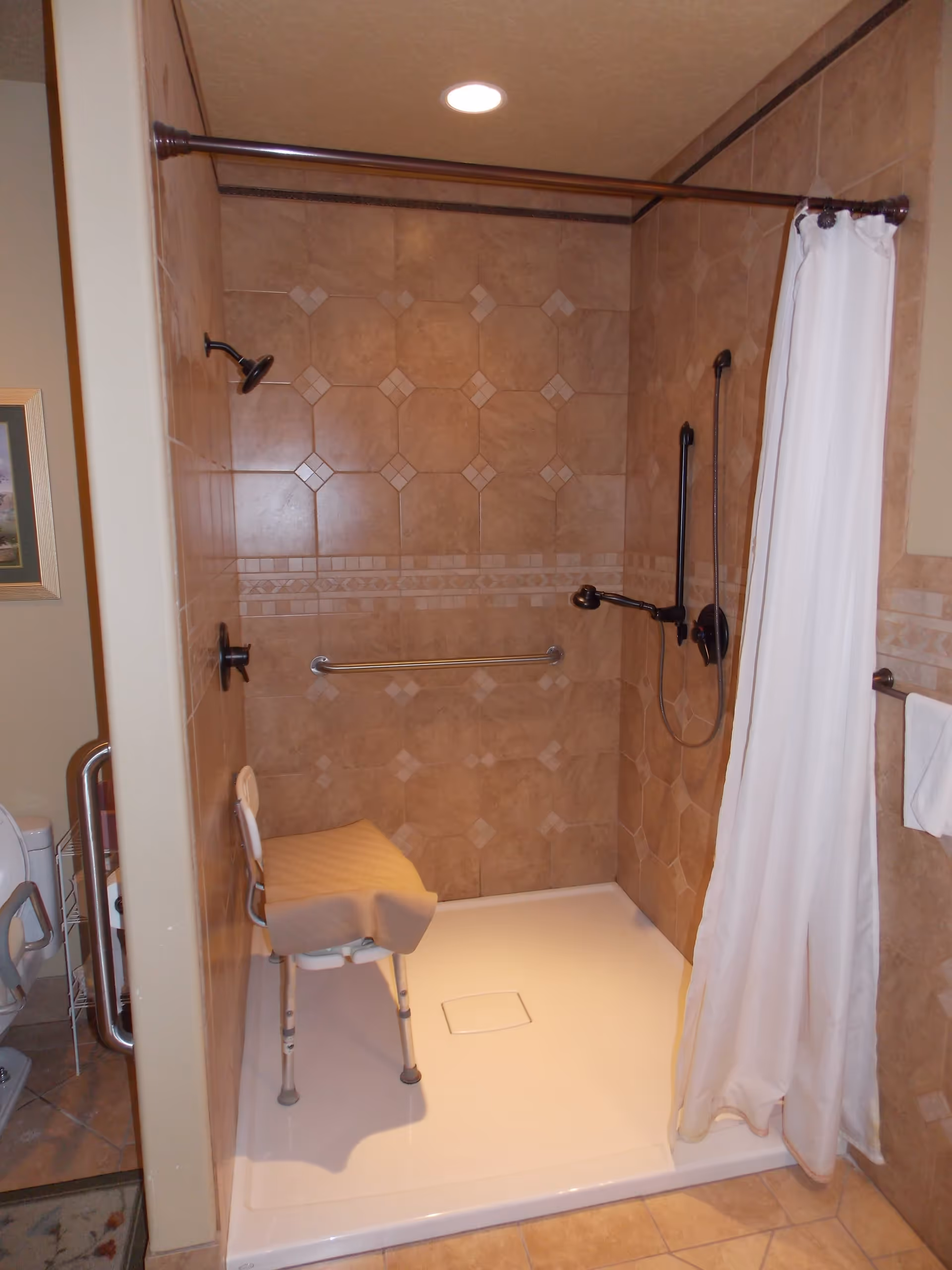 A tiled walk-in shower with a white shower curtain partially drawn to the side. Inside the shower, there is a white shower chair with a beige towel draped over it, a handheld showerhead, and a grab bar on the wall. The tiles are light brown with decorative patterns. The shower floor is white and slightly raised.