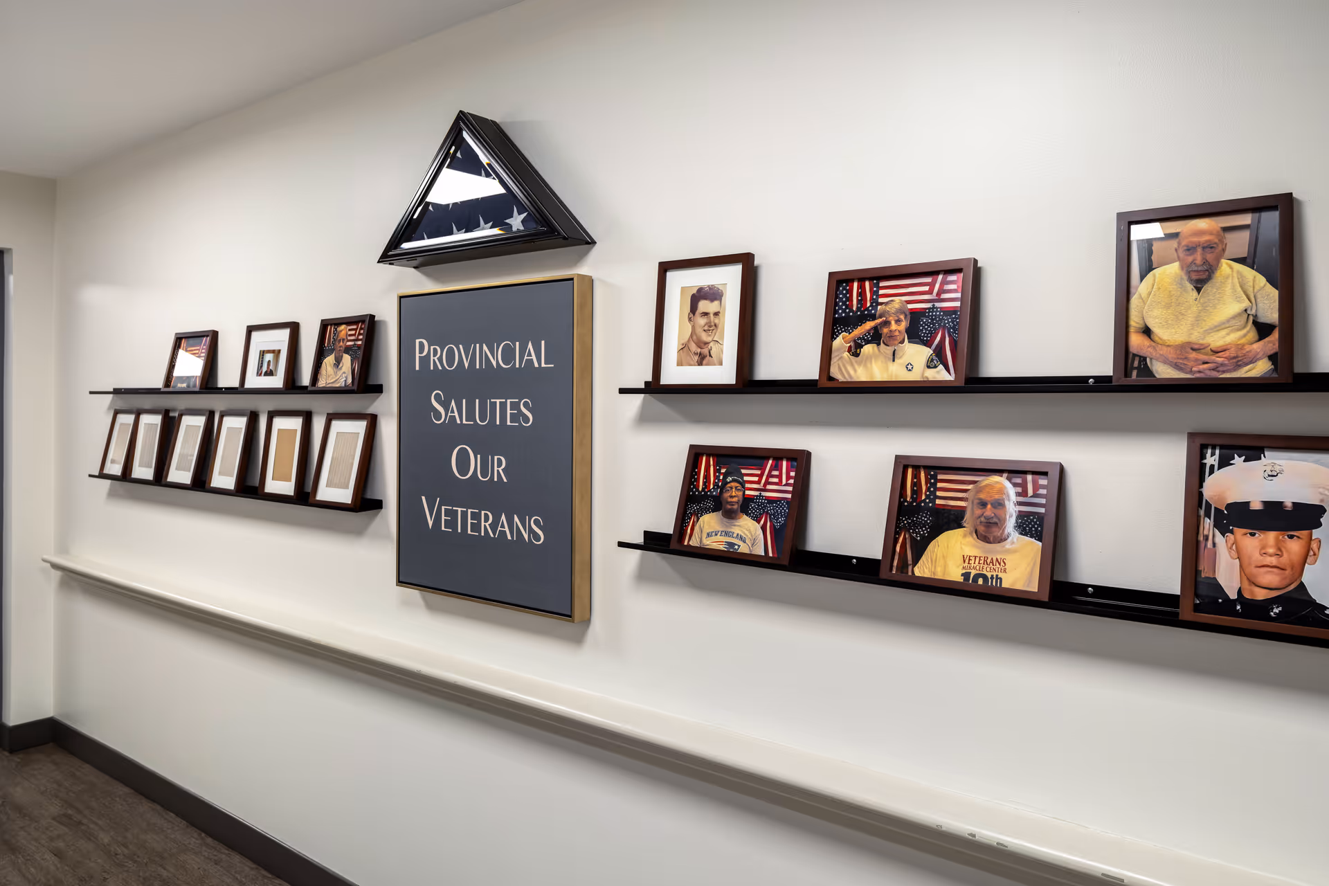 A hallway wall display honoring veterans with framed photographs of veterans, an American flag folded in a triangular case, and a sign that reads 'Provincial Salutes Our Veterans'.