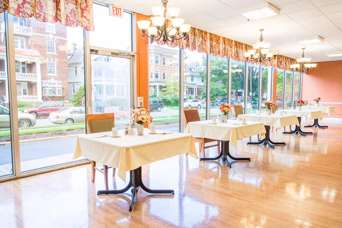 Bright dining room with small tables draped in white tablecloths and floral centerpieces along floor-to-ceiling windows.