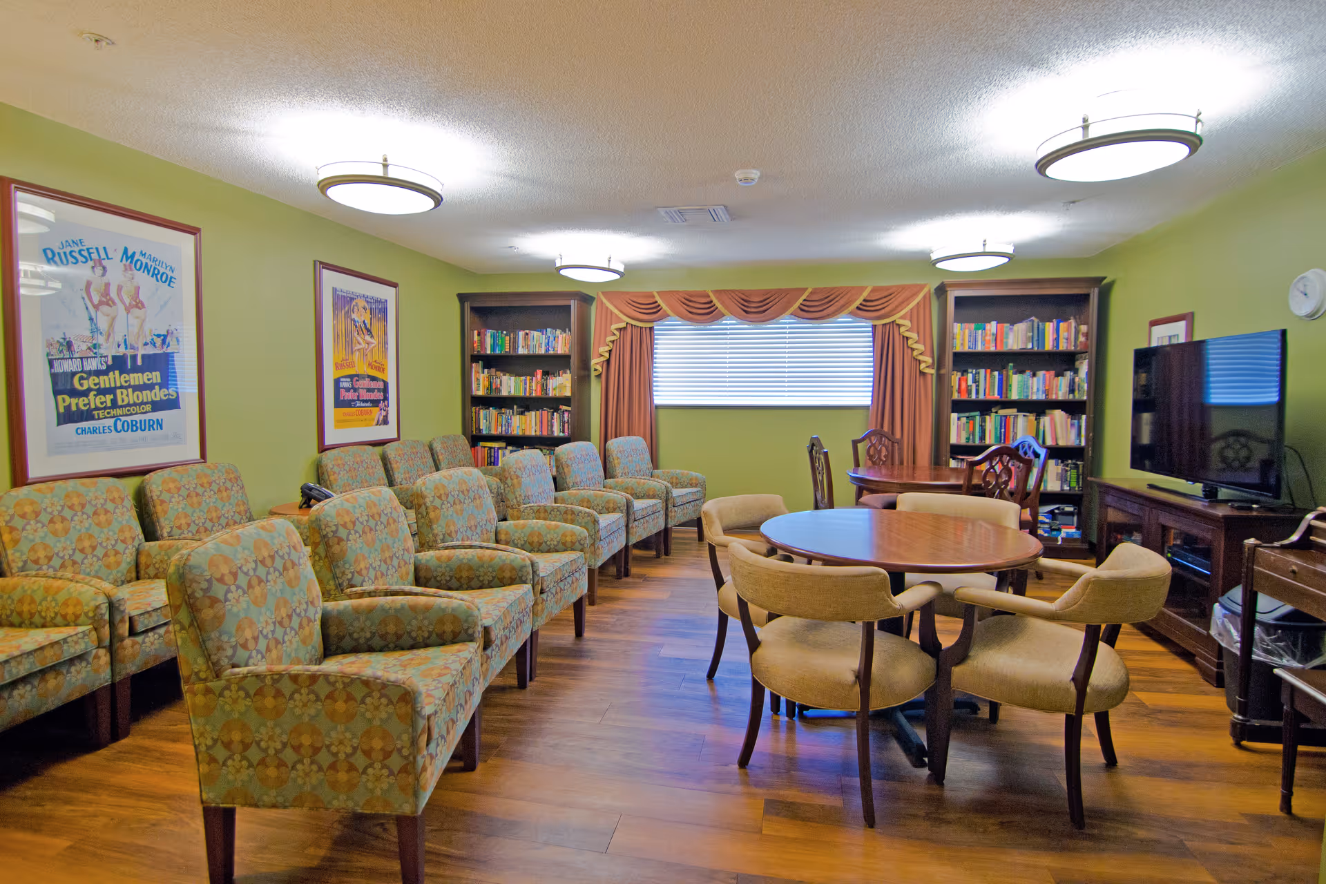 A cozy common room with green walls featuring two framed vintage movie posters. The room has a row of patterned armchairs along one wall and two wooden bookshelves filled with books. In the center, there are two round wooden tables surrounded by beige upholstered chairs. A flat-screen TV sits on a wooden stand against the right wall, and a window with blinds and decorative curtains is at the back.
