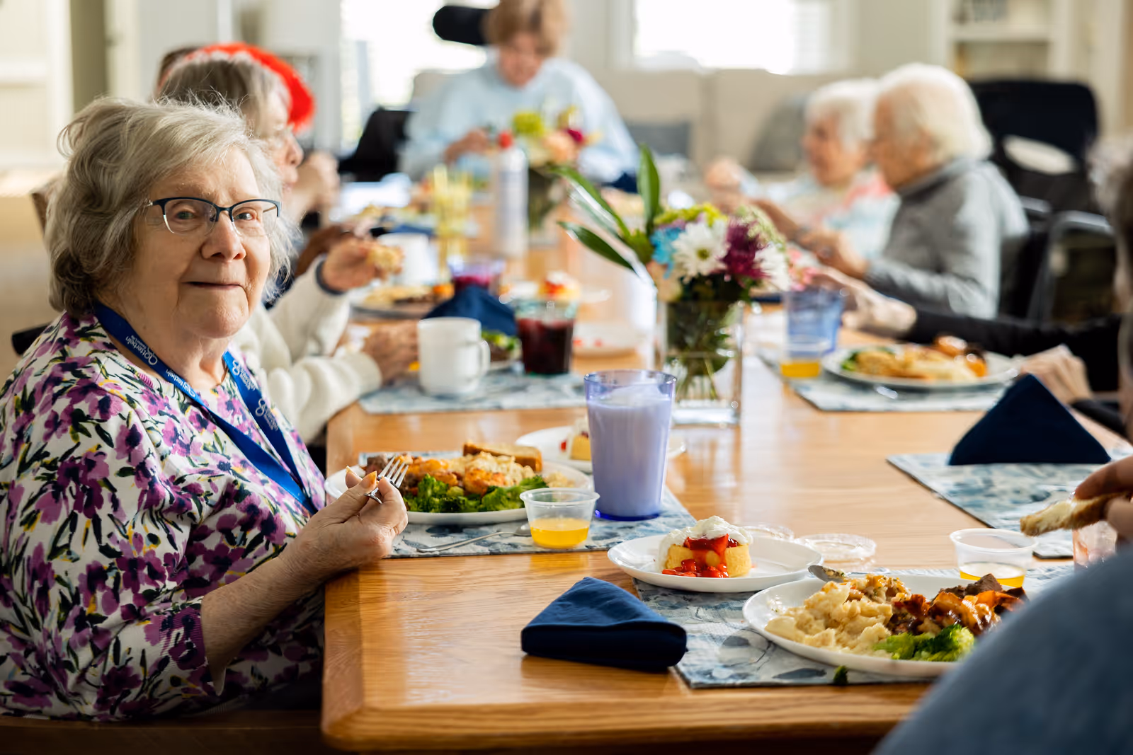 A group of elderly people sitting around a long wooden dining table enjoying a meal together. The table is set with plates of food, drinks, and a vase with colorful flowers in the center. One elderly woman in the foreground is looking at the camera while holding a fork.