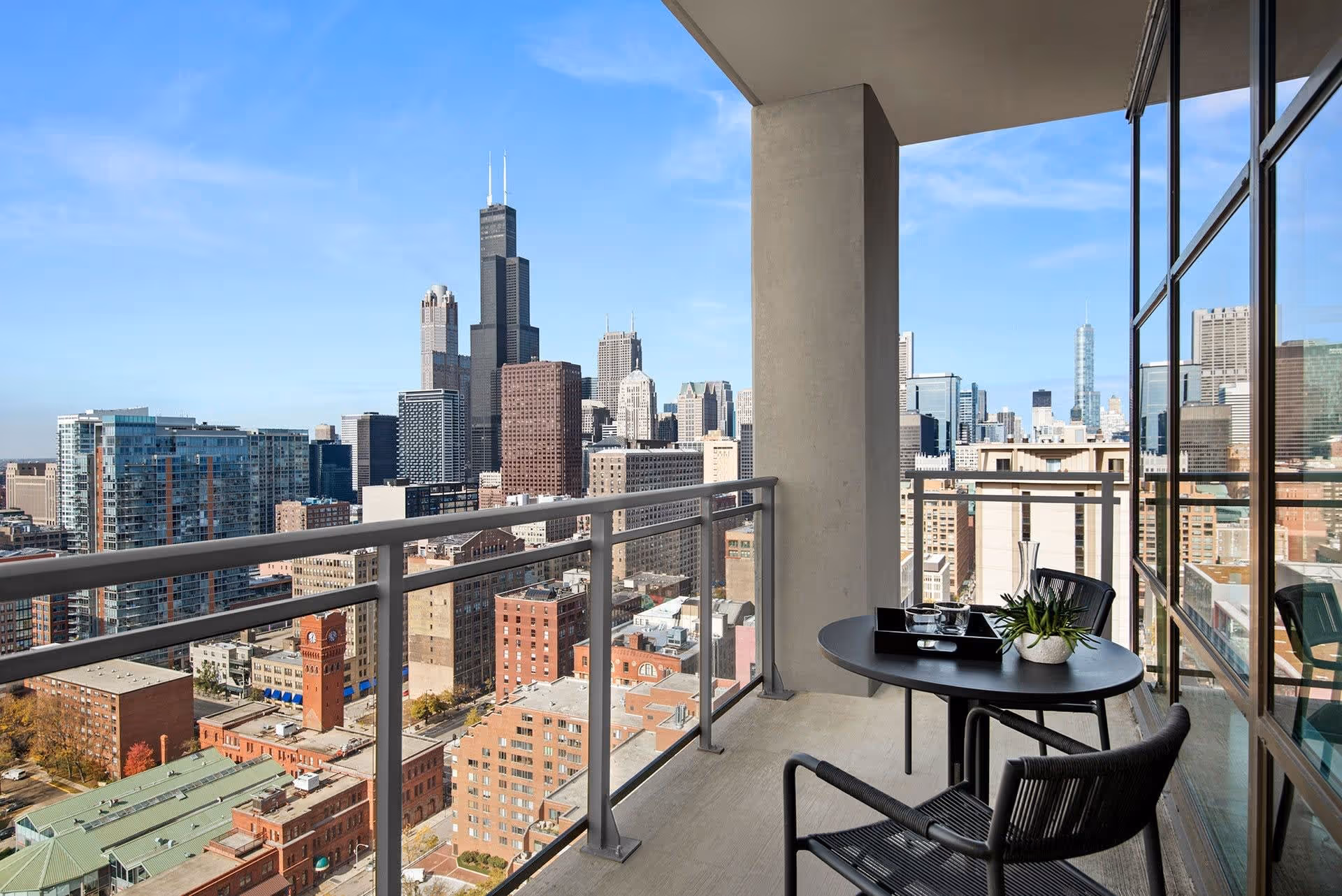 Balcony with two black chairs and a small round table with a plant and glassware, overlooking a city skyline with tall buildings under a clear blue sky.