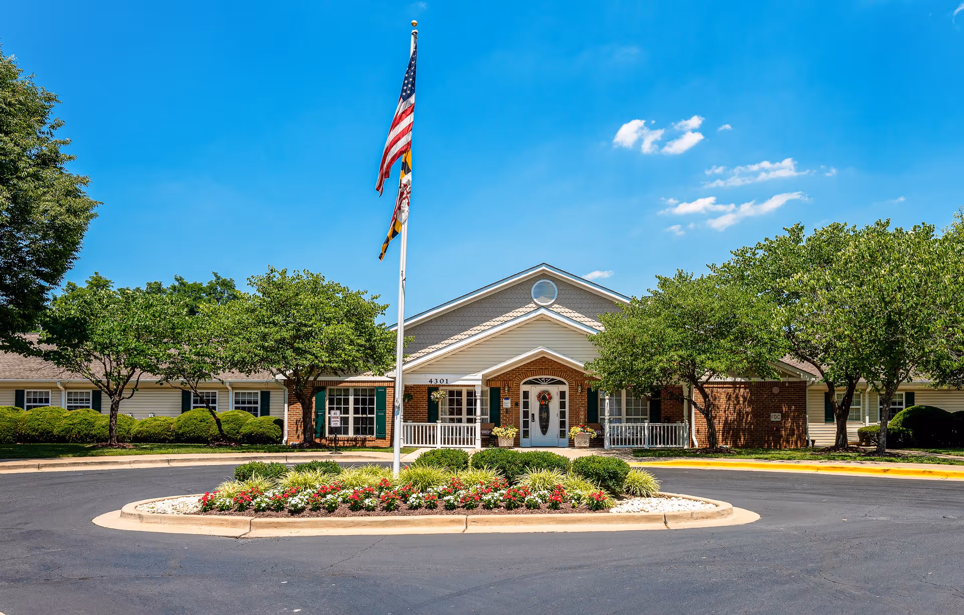Front exterior view of Arden Courts - ProMedica Memory Care Community (Kensington) building with a circular driveway, landscaped flower bed, American flag and Maryland state flag on a flagpole, surrounded by green trees under a clear blue sky.