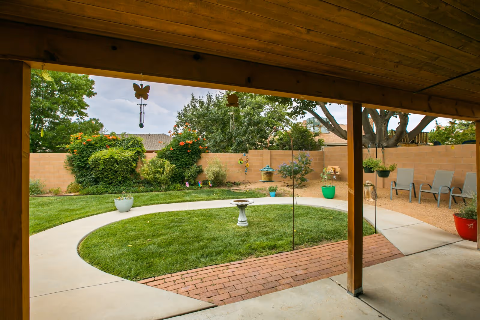 View of a covered patio looking out onto a circular grassy area with a birdbath in the center, surrounded by a concrete walkway. The garden area has various potted plants, hanging planters, and chairs along a brick wall with trees and shrubs in the background.