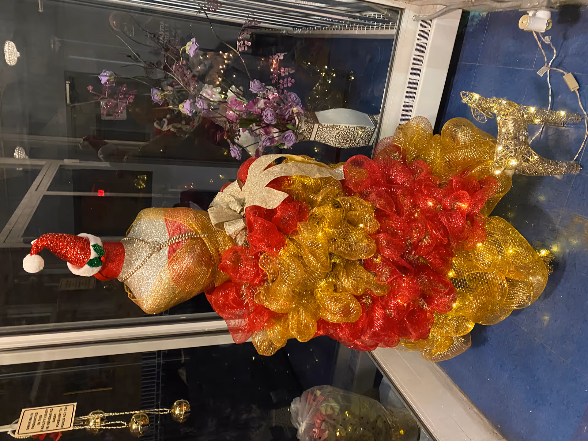 A decorative mannequin dressed in a festive red and gold dress with a Santa hat, adorned with a pearl necklace, standing on a blue floor inside a glass enclosure. Next to the mannequin is a small illuminated reindeer figure. Behind the mannequin, there is a vase with purple flowers and some holiday decorations reflected in the glass.
