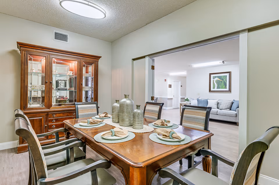 A dining room with a wooden table set for four with plates, napkins, and decorative vases in the center. There is a wooden china cabinet with glass doors displaying dishes and decor. The room opens into a living area with a gray sofa and a framed bird artwork on the wall.