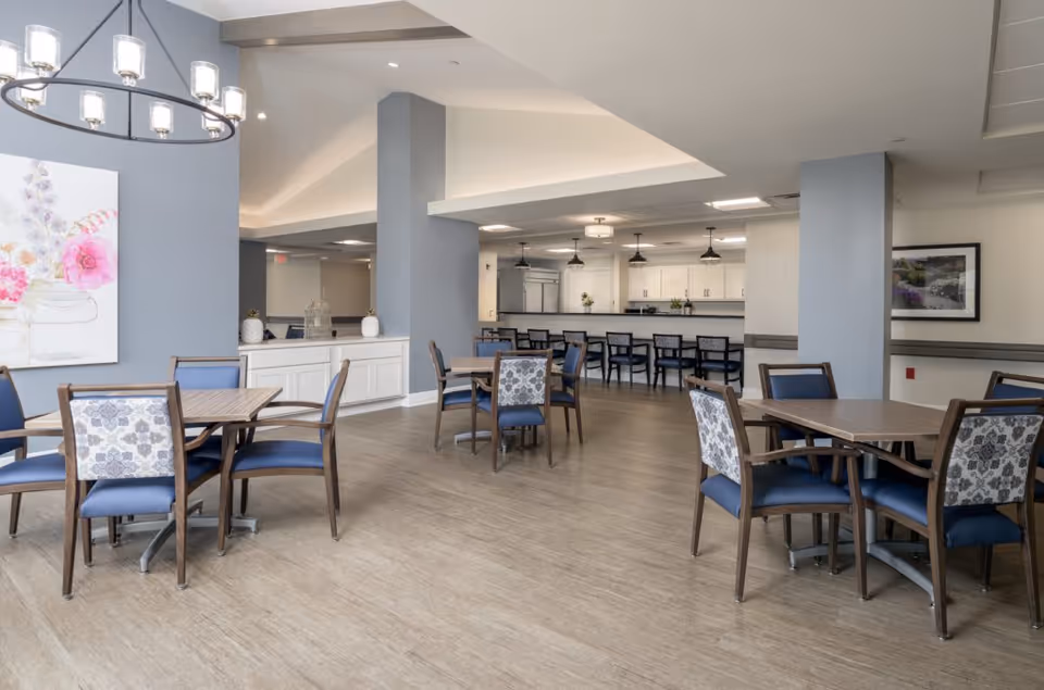 Spacious dining area with multiple wooden tables and blue upholstered chairs, a service counter and pendant lights in a senior living facility.