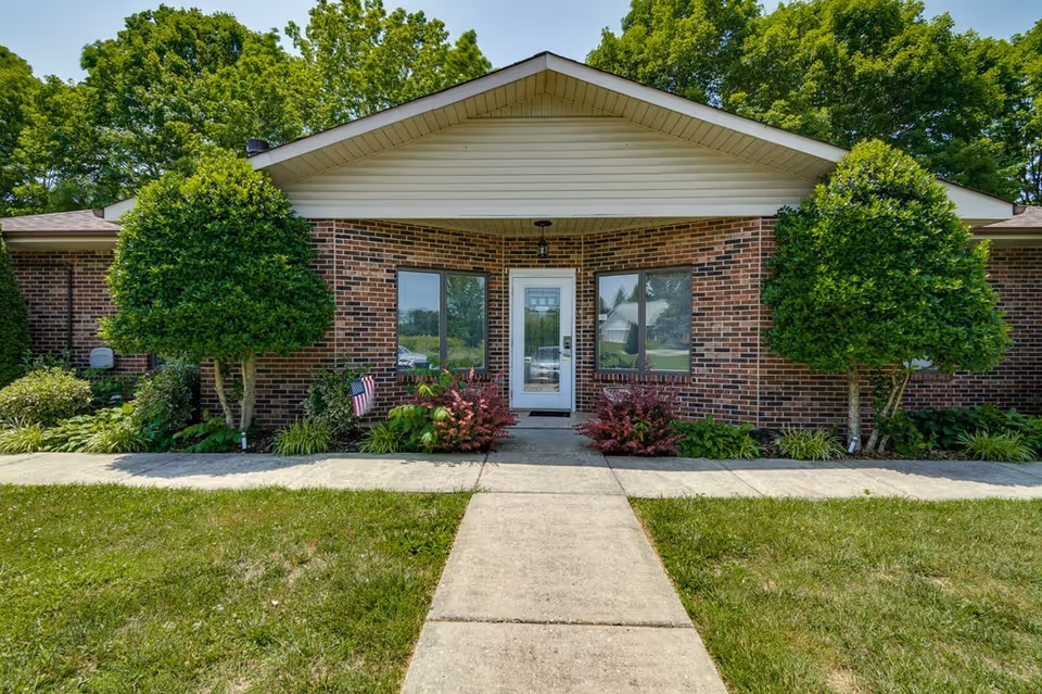 Front exterior view of a single-story brick building with a white door and two large windows on either side. The entrance is flanked by two neatly trimmed trees and various shrubs, with a concrete walkway leading up to the door. The building is surrounded by green grass and trees in the background under a clear blue sky.