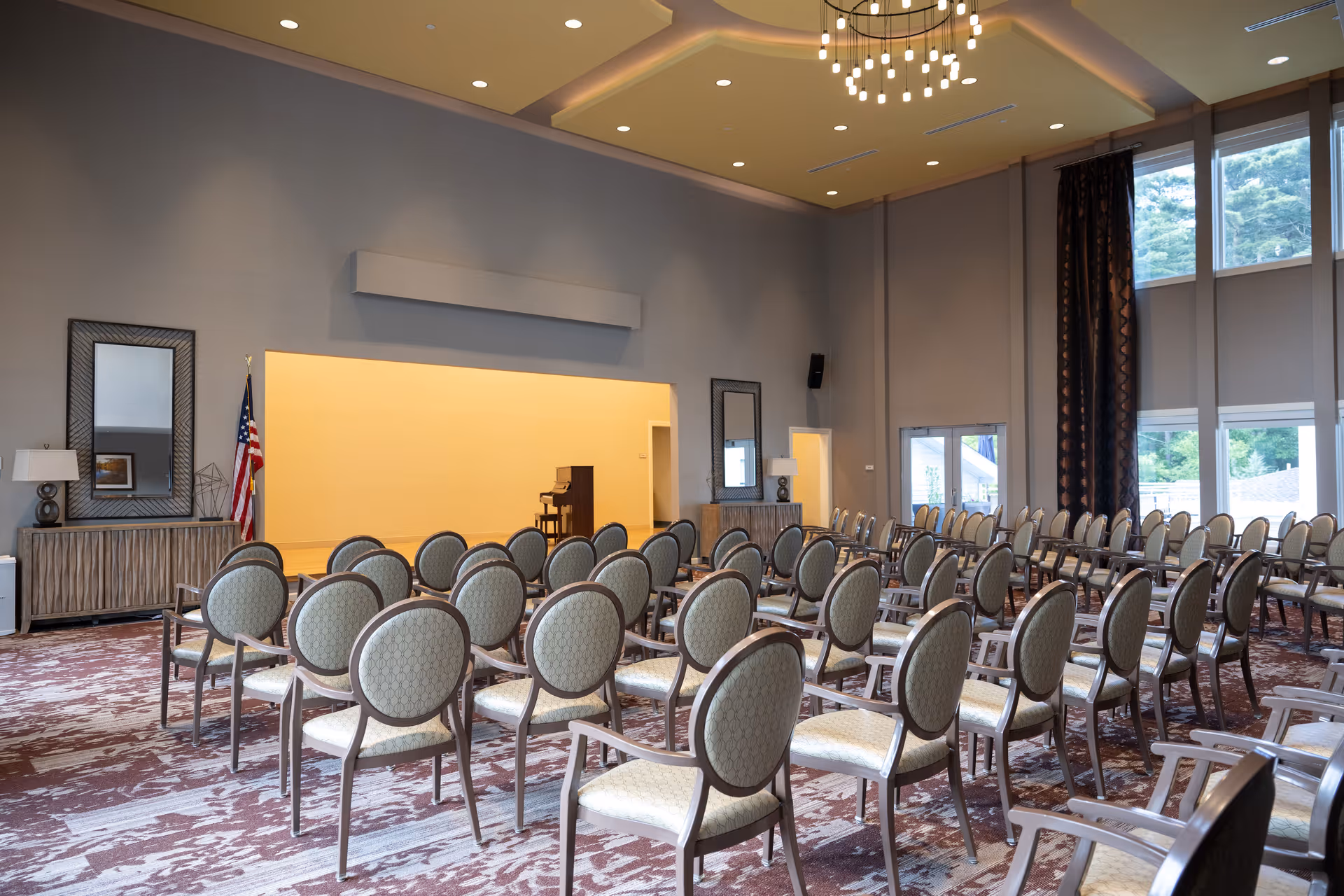 A large room with rows of upholstered chairs arranged facing a small stage area with a piano. The room has high ceilings with recessed lighting, large windows with dark curtains, and neutral-colored walls. There are two mirrors and lamps on side tables along the walls, and an American flag is positioned near the stage.