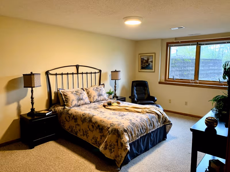 A bedroom with a double bed featuring floral bedding, two bedside tables each with a lamp, a black leather chair near a window, a framed picture on the wall, and a dresser with decorative items. The room has beige walls and carpeted floor.