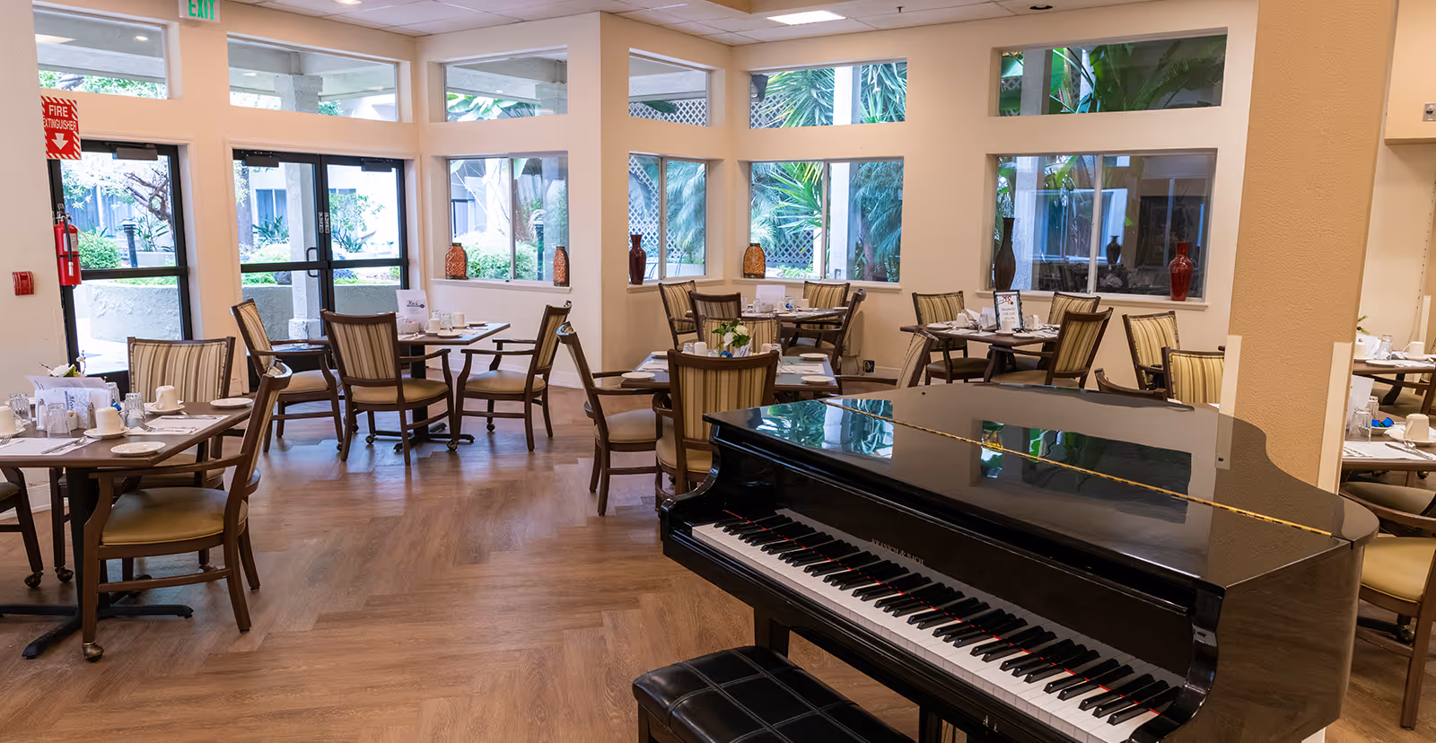 A bright dining room with multiple tables and chairs set for a meal, large windows letting in natural light, and a black grand piano in the foreground.