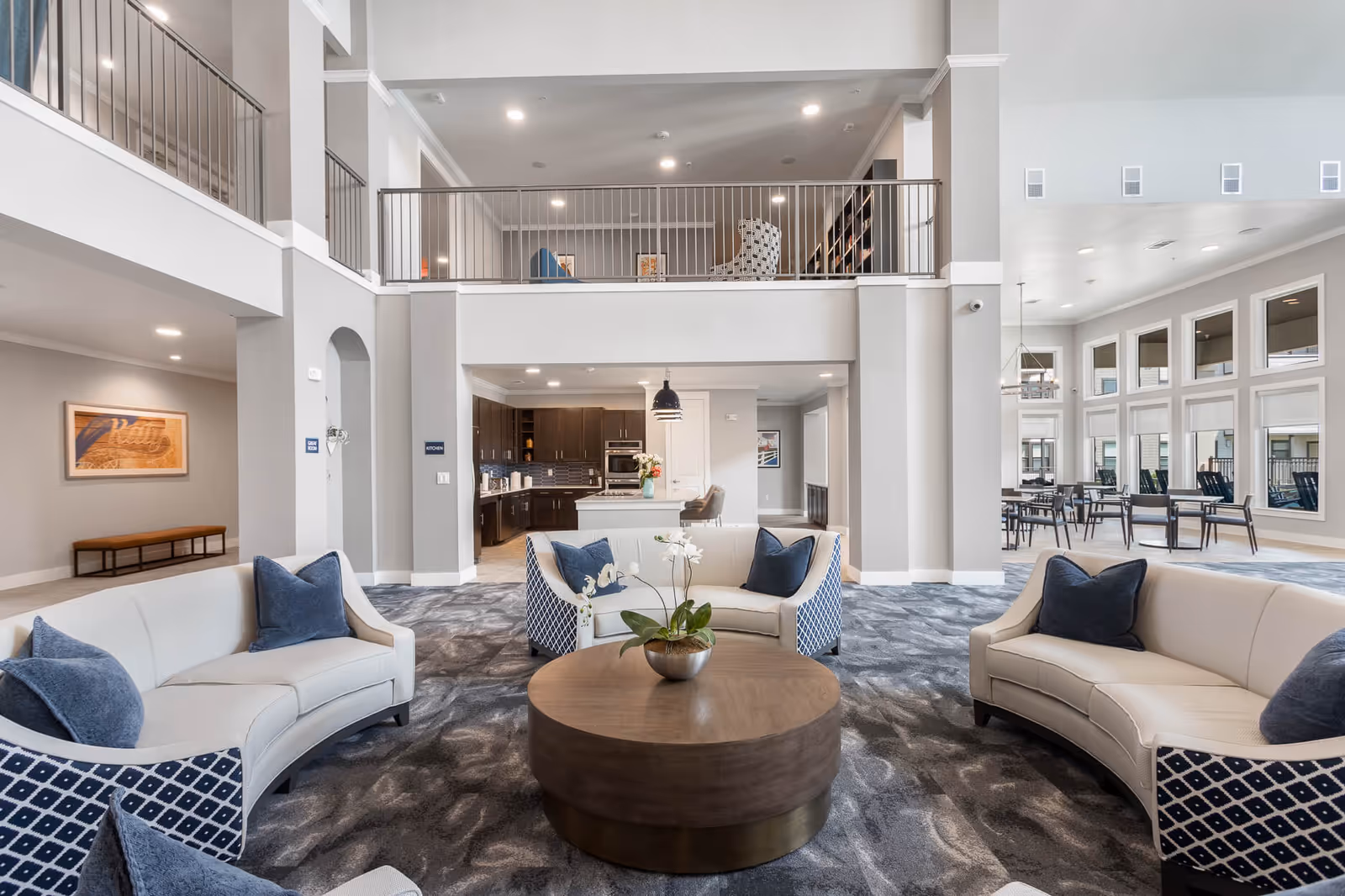 Spacious and modern senior living facility common area with curved sofas featuring blue and white patterned upholstery and blue pillows arranged around a large round wooden coffee table with a potted orchid. The area has a high ceiling with a second-floor balcony railing overlooking the space. In the background, there is a kitchen area with dark cabinets and a dining area with multiple tables and chairs near large windows.