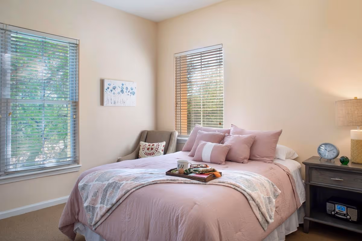 A cozy bedroom with a pink-made bed topped with multiple pillows, an armchair by two blinds-covered windows, and a nightstand with a lamp and clock.