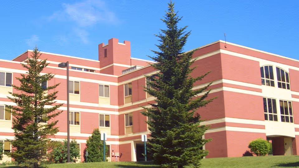 Exterior view of a multi-story brick building with white horizontal stripes, surrounded by green grass and tall evergreen trees under a clear blue sky.