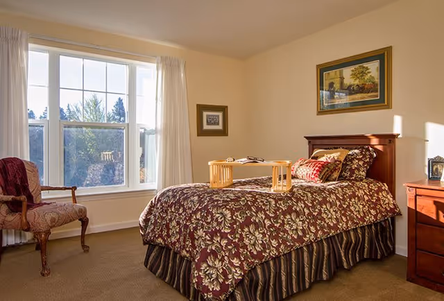 A cozy bedroom with a single bed covered in a floral patterned bedspread and multiple pillows. A wooden tray with a book and glasses sits on the bed. There is a wooden chair with an upholstered seat and backrest near a large window with white curtains. The walls are light-colored and decorated with framed artwork. A wooden nightstand with a lamp is beside the bed.