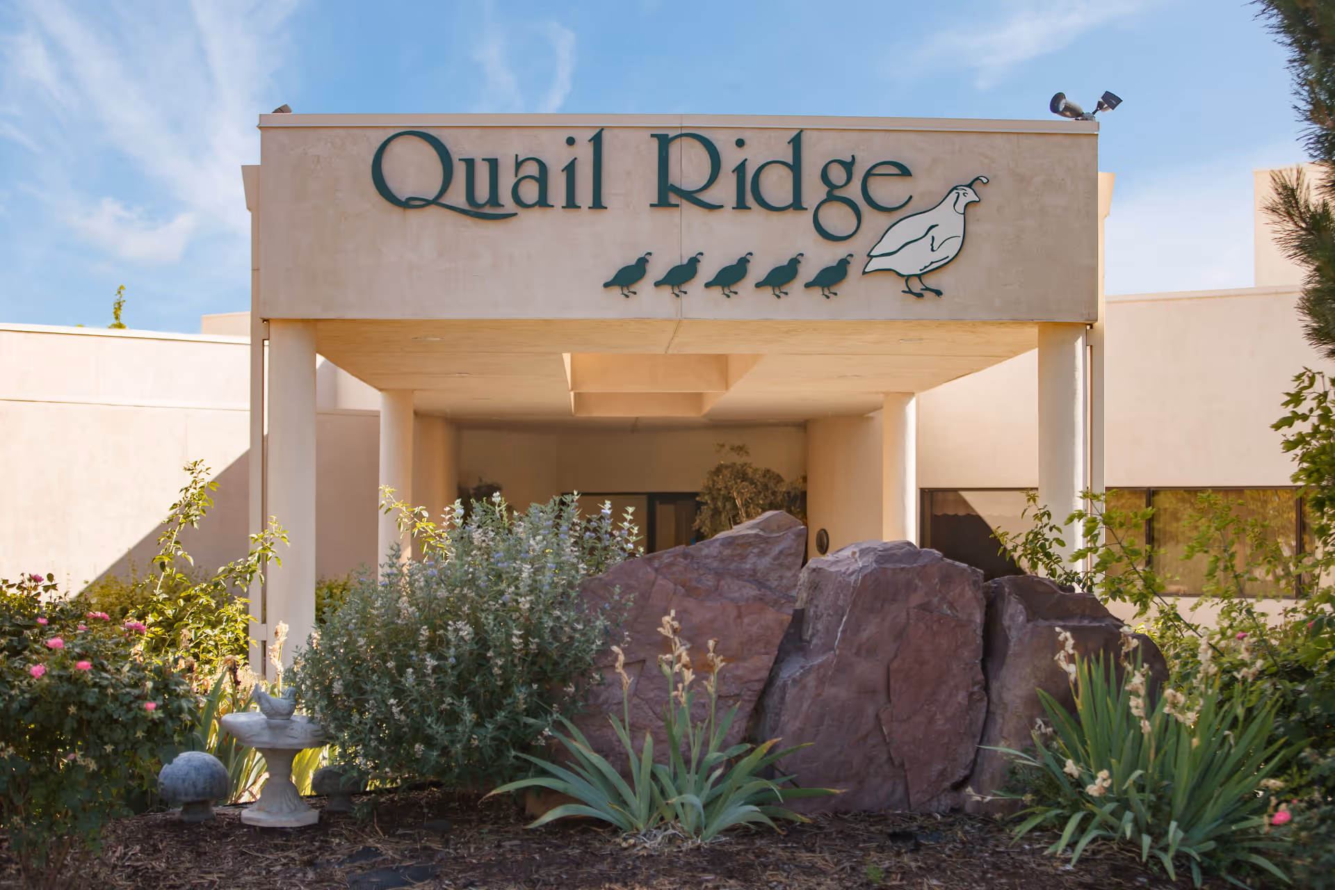 Entrance of Quail Ridge facility with a beige building facade, large rocks, green shrubs, and plants in the foreground under a blue sky.