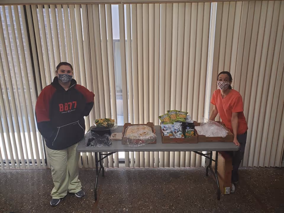 Two people wearing face masks stand on either side of a folding table with various food items, including salad, packaged snacks, and covered trays, in front of vertical blinds.