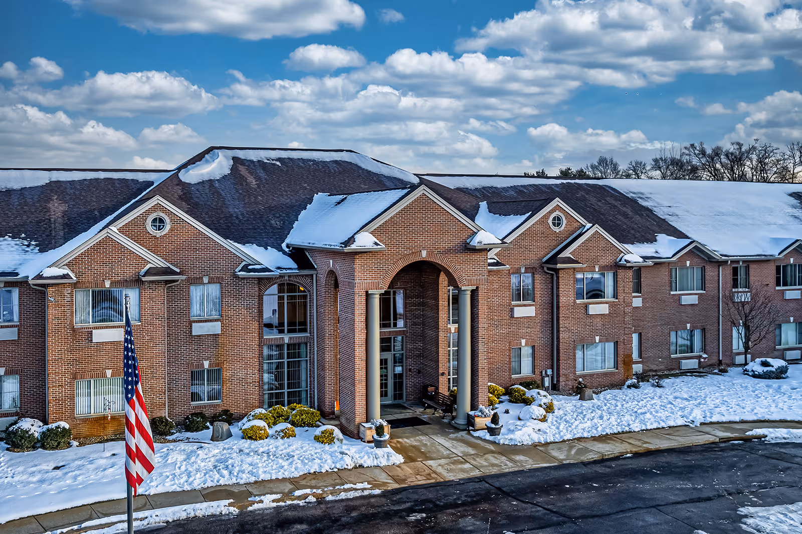 Front exterior view of a two-story brick building with snow on the roof and ground. The building has large windows, a covered entrance with columns, and an American flag in front. The sky is partly cloudy.