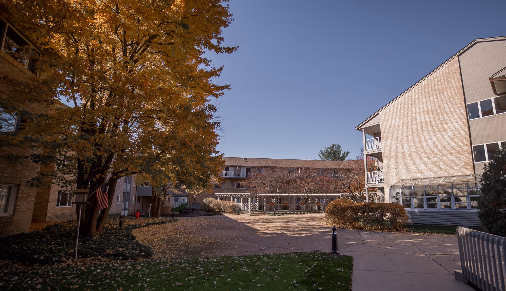 Outdoor courtyard area of a retirement community with autumn trees, a paved walkway, and multi-story brick buildings surrounding the space under a clear blue sky.