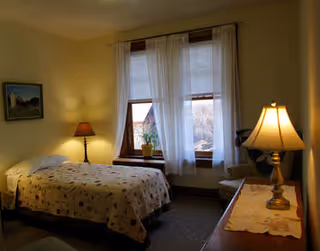 A cozy bedroom with a single bed covered in a floral quilt, a bedside table with a lamp, a window with sheer white curtains, and a dresser with another lamp and a decorative cloth.
