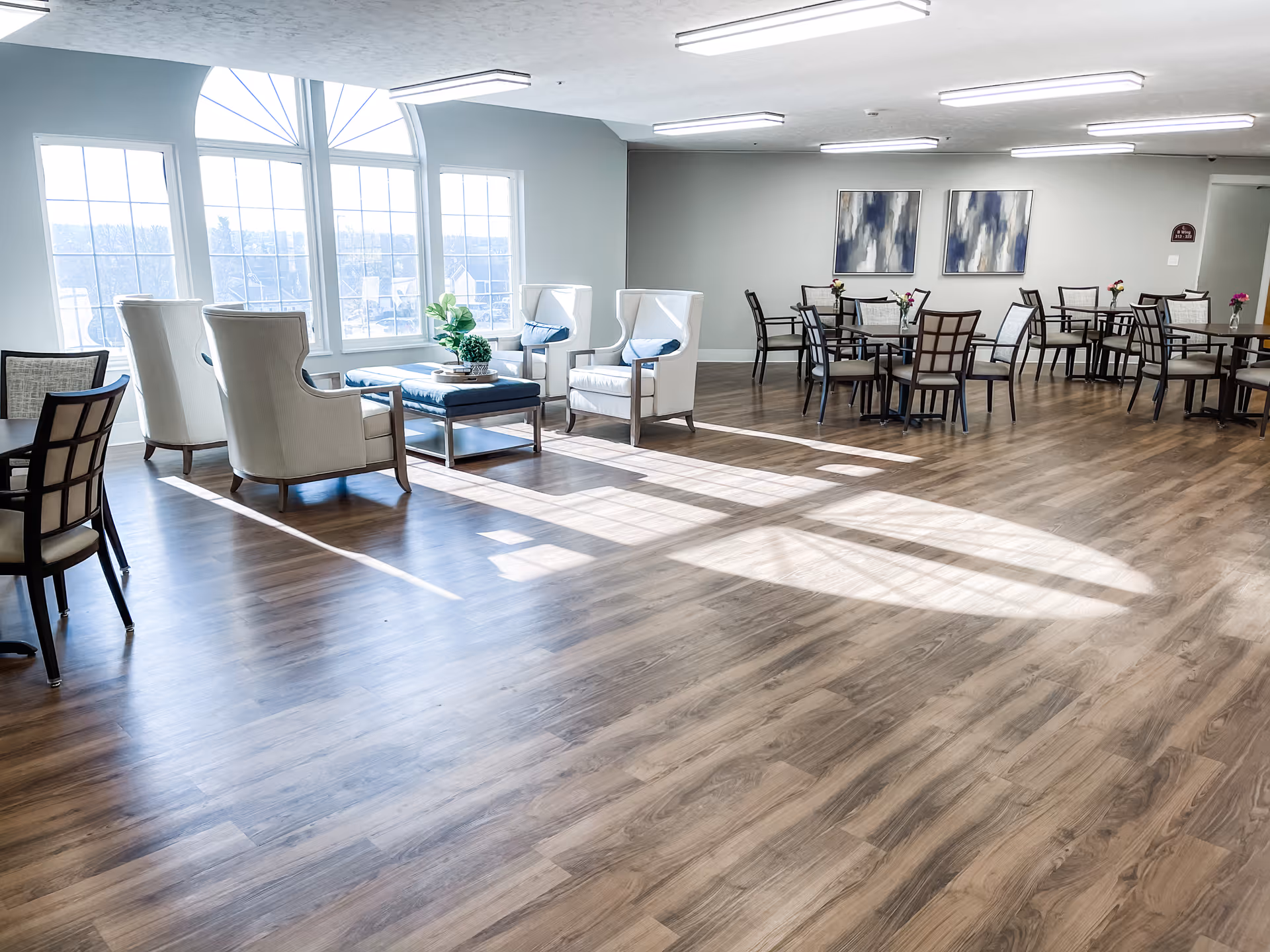 A bright and spacious common area with large arched windows letting in natural light. The room features several white armchairs with blue cushions arranged around a coffee table, and multiple dining tables with chairs. The floor is wood, and the walls are painted light gray with two abstract paintings hanging on one wall.