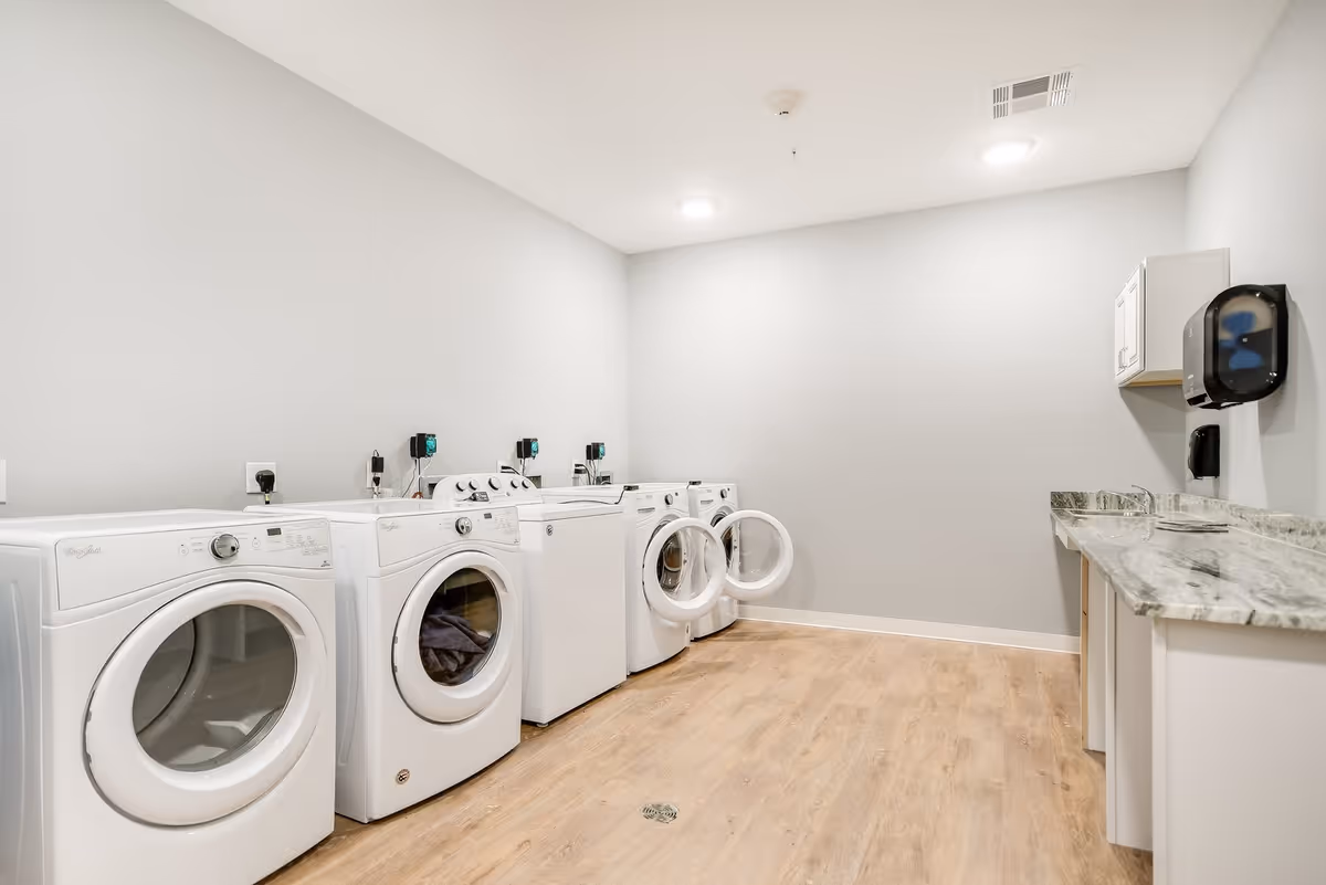 A clean and modern laundry room with four white washing machines and dryers lined up against a light gray wall. The floor is wood-style laminate, and there is a countertop with a marble pattern on the right side along with a wall-mounted paper towel dispenser and soap dispenser.